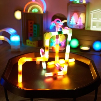Luzes LED coloridas em estrutura tubular sobre mesa preta com brinquedos coloridos ao fundo