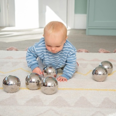 Bebé com camisola azul riscada e bolas metálicas prateadas no tapete.