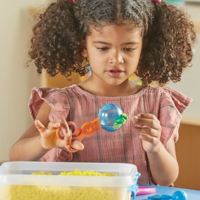 Criança com vestido rosa a brincar com utensílio e bola azul em mesa com recipiente plástico