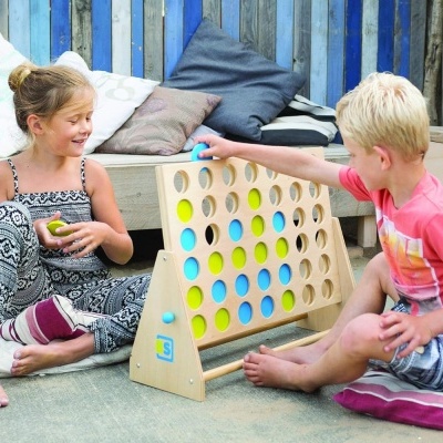 Duas crianças a jogar um jogo de tabuleiro em madeira com fichas azuis e amarelas num ambiente acolhedor com sofás e almofadas.