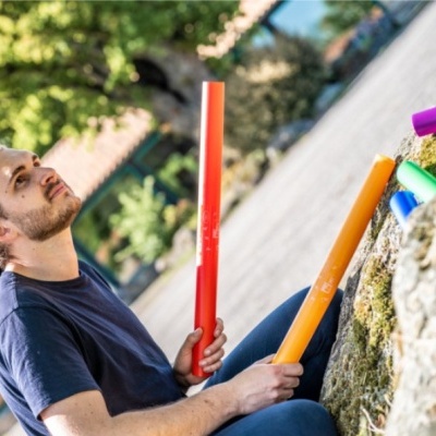Homem sentado segurando tubos coloridos de plástico com texto branco, ao lado de rocha com outros tubos coloridos