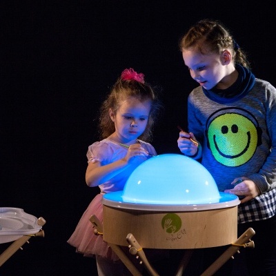 Duas crianças interagem com mesa redonda de madeira com cúpula azul luminosa num ambiente escuro