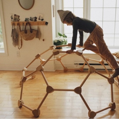 Estrutura de brincar infantil em madeira clara em sala com chão e paredes claras, janela, espelho redondo e criança a brincar