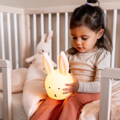 Criança sentada na cama a segurar um candeeiro em forma de coelho amarelo com orelhas grandes