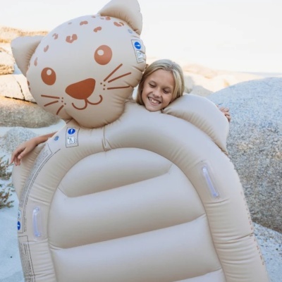 Boia insuflável infantil formato gato bege com detalhes castanhos e etiquetas azuis, exterior rochoso