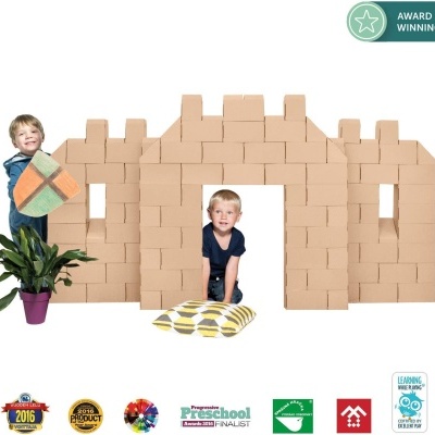 Crianças brincando com blocos de construção que formam uma casa, segurando almofadas coloridas e uma planta em vaso.