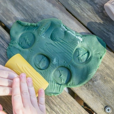 Brinquedo de plastilina verde moldada com rolo amarelo, mãos infantis, sobre madeira, tubos brancos ao lado