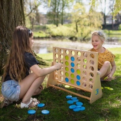 Jogo de tabuleiro gigante de madeira com peças azuis e amarelas ao ar livre junto a árvore.