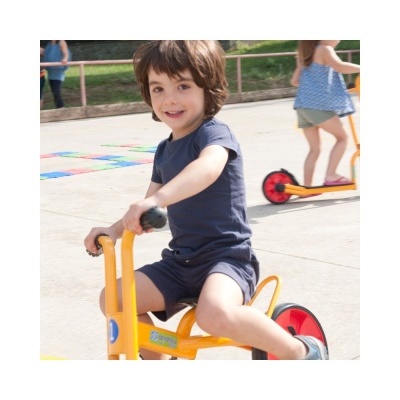 Criança num triciclo amarelo num espaço exterior com outras crianças e um trotinete