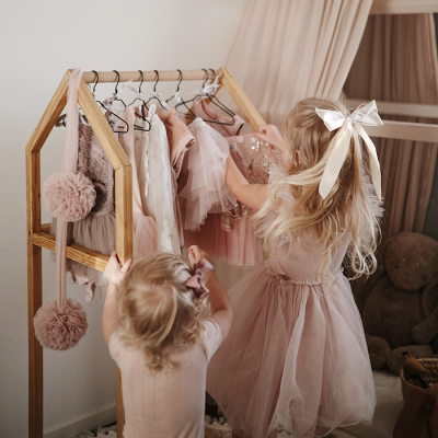 Vestidos de menina rosa pendurados em cabideiro de madeira com duas crianças a olhá-los