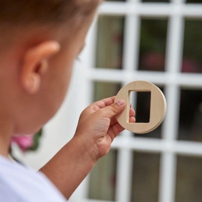 Criança segurando espelho redondo de madeira com buraco retangular próximo a janela branca