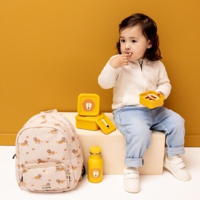 Criança com mochila bege estampada com leões, caixas e garrafa amarelas com rosto de leão, sentada em banco contra parede laranja.