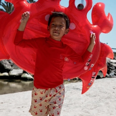 Criança com bóia insuflável vermelho caranguejo na praia com roupa vermelha e calções bege com caranguejos