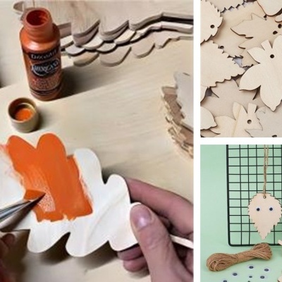 Mãos a pintar figura de madeira com tinta laranja, perto de tintas e várias figuras de madeira em formato de folhas e animais.