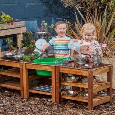 Mesa de brincar de madeira com acessórios de cozinha e duas crianças a brincar num jardim