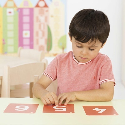 Menino de camisa rosa com cartas numeradas vermelhas à frente, sentado numa mesa branca em ambiente interior colorido