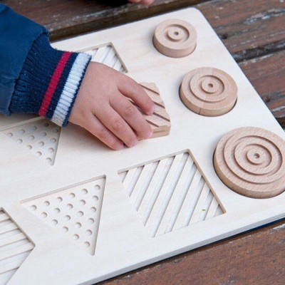 Painel sensorial de madeira com formas geométricas e textura, mão de criança a segurar peça.