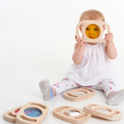 Bebé com túnica branca segura moldura de madeira com vidro amarelo e está sentado junto a outras molduras coloridas.