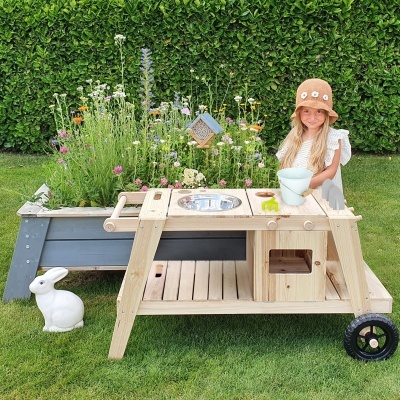 Mesa de madeira clara com rodas e brinquedos, junto a bancada azul com flores, em relva
