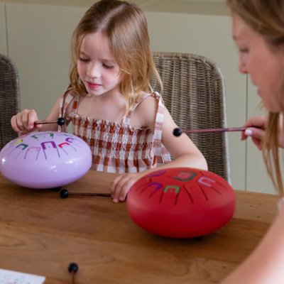 Duas crianças a tocar tambores circulares de aço pintados em lilás e vermelho numa mesa de madeira.