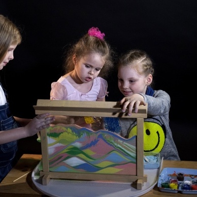 Três crianças manipulam caixa de areia colorida com padrões sobre mesa branca e fundo preto