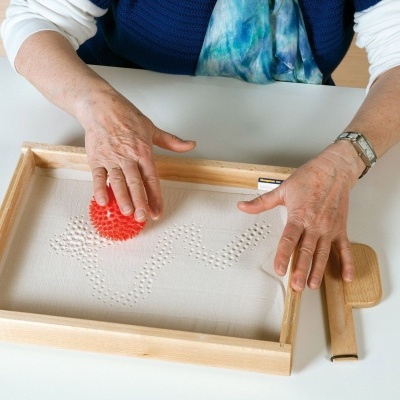 Mãos femininas manipulam moldura de madeira com tabuleiro branco e pequenas esferas