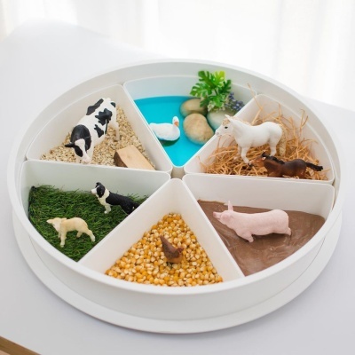 Tabuleiro redondo com compartimentos de diferentes materiais e figuras de animais da fazenda.