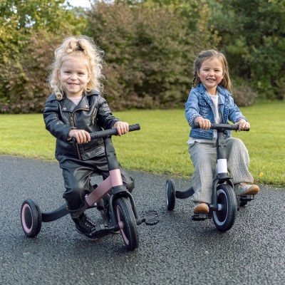 Duas crianças em bicicletas de equilíbrio tricolor numa estrada com relva ao fundo