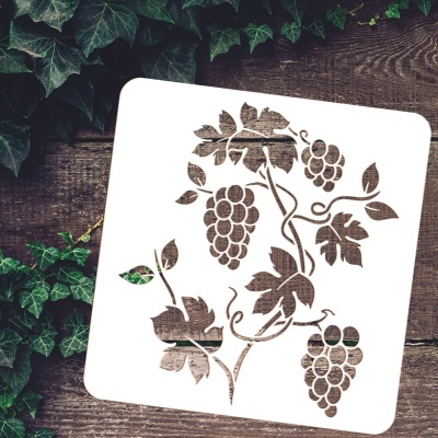 Stencil branco com desenho de cachos de uvas e folhas colocado sobre madeira com folhagem verde.