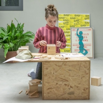 Criança a brincar com blocos de madeira numa mesa de aglomerado com planta e quadros ao fundo