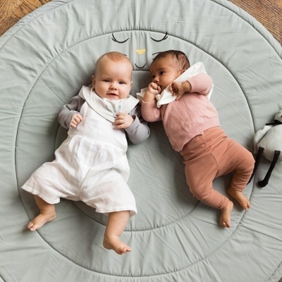 Dois bebés deitados num tapete redondo verde claro com padrão de rosto de animal
