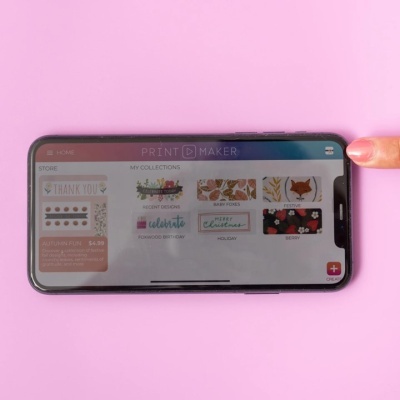 Smartphone com ecrã da aplicação PRINT MAKER e dedo a tocar o canto superior direito sobre fundo rosa