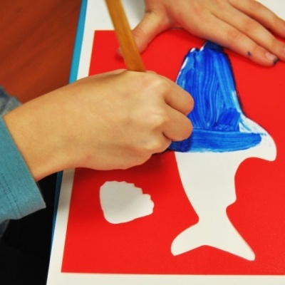 Criança a colorir um peixe recortado em papel com lápis de cor azul sobre fundo vermelho.