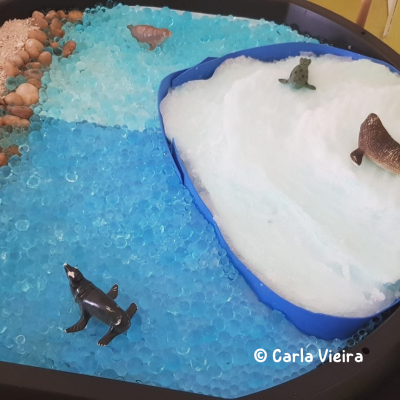 Mesa sensorial com gel azul, ovos de plástico e figuras de animais marinhos.