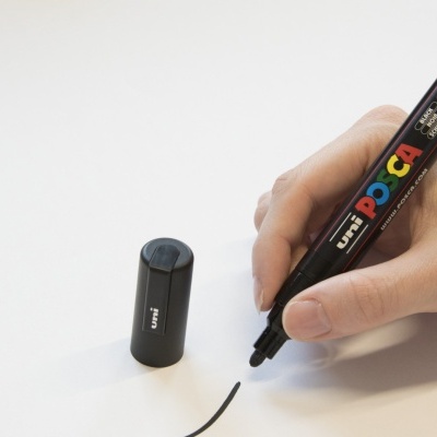 Marcador preto POSCA com tampa ao lado e mão a escrever numa folha branca