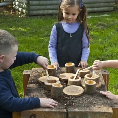 Crianças a brincar com utensílios de cozinha de madeira ao ar livre numa mesa rústica.