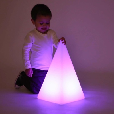 Criança ajoelhada com luminária piramidal cor-de-rosa em ambiente com pouca luz