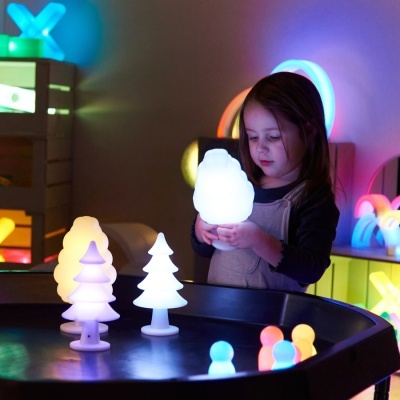 Menina a segurar uma luz branca com outras luzes coloridas em forma de árvores numa mesa preta.