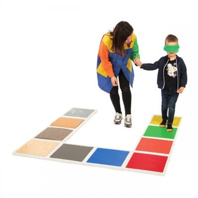 Tapetes sensoriais quadrados coloridos e texturizados no chão com uma criança vendada e uma mulher a segurá-la pela mão