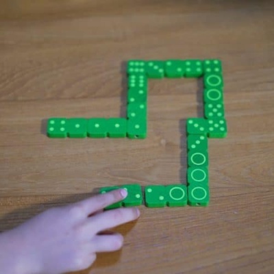 Peças de dominó verdes com pontos brancos dispostas em L sobre mesa de madeira com mão a mexer
