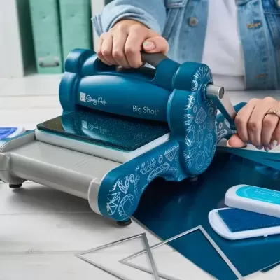 Máquina de corte azul escura Big Shot Sizzix com manivela sobre mesa branca e moldes metálicos