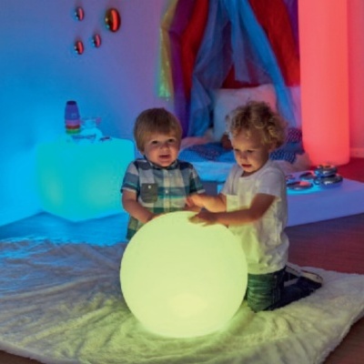 Crianças brincam com bola luminosa verde num tapete branco com iluminação colorida ao fundo