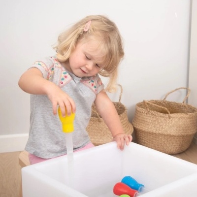 Menina loira a brincar com brinquedos coloridos num recipiente branco junto a cestos de fibra natural