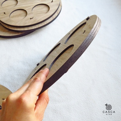 Peça redonda de madeira com recortes ovais segurada por mão, fundo branco e logotipo CASCA VIVA.