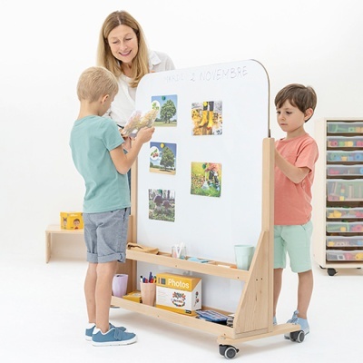 Quadro branco duplo de madeira com prateleira e rodas num ambiente branco com duas crianças e uma mulher