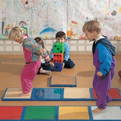 Crianças brincam com tapetes táteis e cubos de espuma numa sala com desenhos na parede