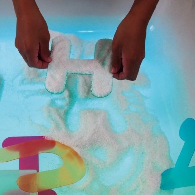 Mãos segurando letra H branca sobre grânulos com letras plásticas coloridas