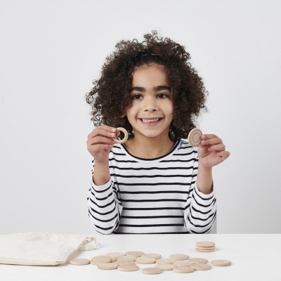 Criança com t-shirt de riscas segurando peças redondas de madeira, com peças e saco de pano sobre a mesa