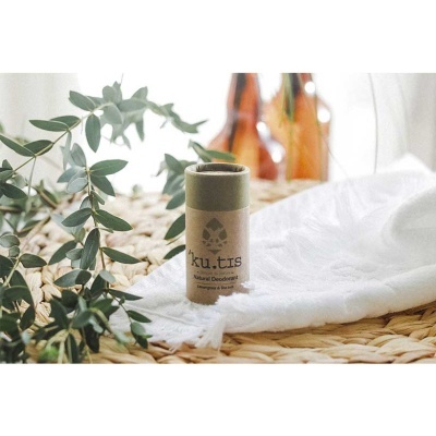 Desodorizante sólido ku.tis em embalagem cilíndrica castanha, pano branco e folhas verdes