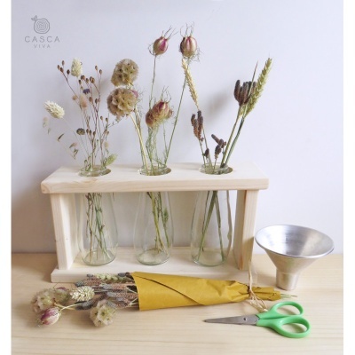 Arranjo de flores secas em frascos de vidro com suporte de madeira, tesoura verde, funil metálico e ramo de flores secas em papel amarelo